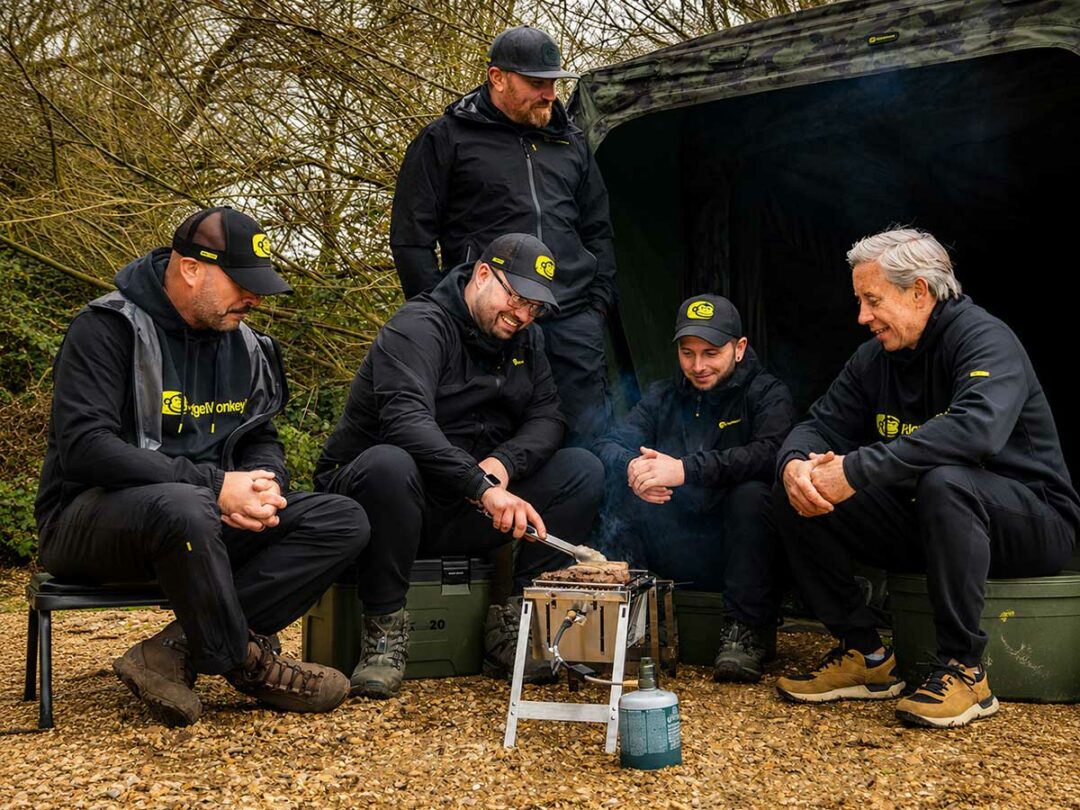 Karpfenangler in moderner RidgeMonkey Angelbekleidung bei einer entspannten Session am Wasser mit Outdoor-Setup und Grill.