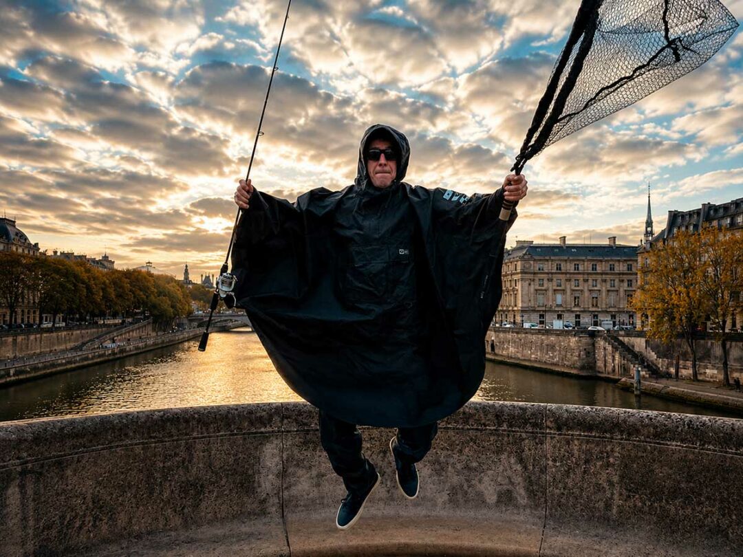Angler in schwarzer Salty Crew Regenbekleidung mit Kescher und Angelrute auf einer Brücke bei Sonnenuntergang