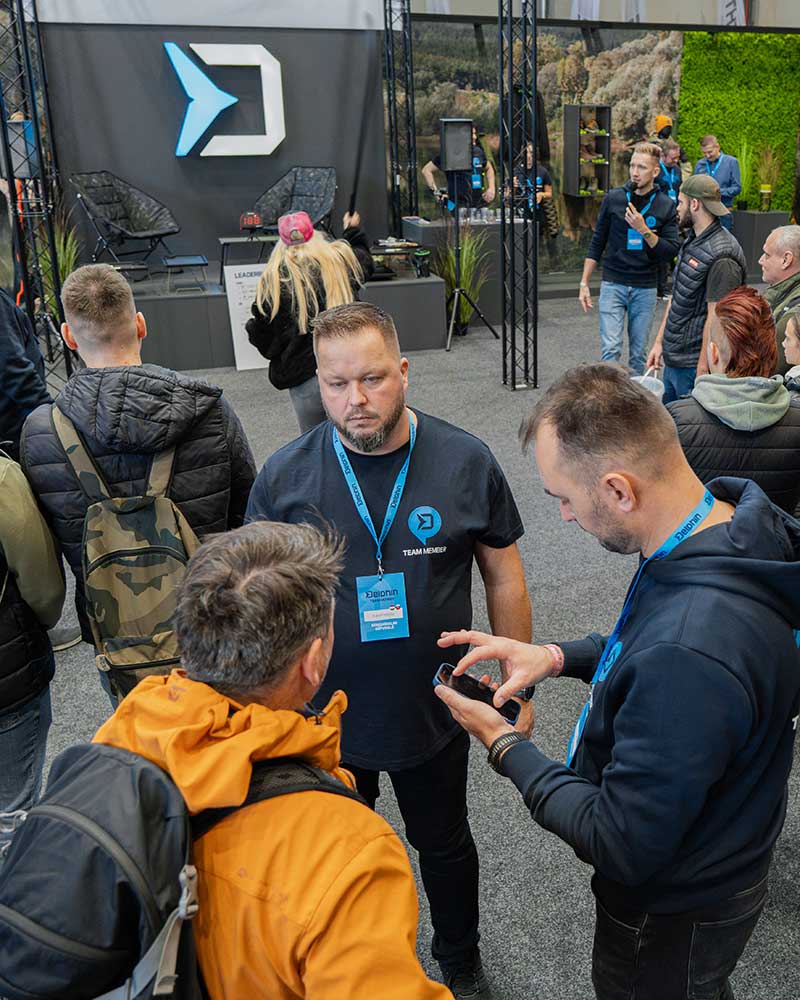 Logo Delphin nad veletržním stánkem, pod ním rybáři porovnávají kaprařské vybavení a vybavení na dravce