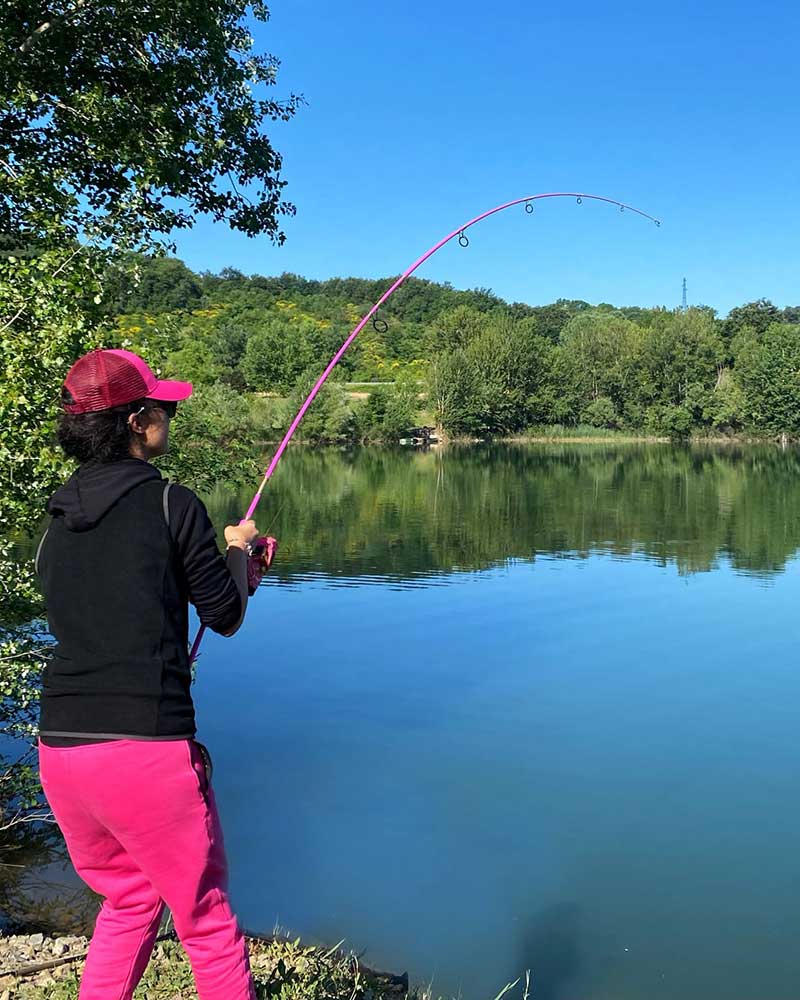 Anglerin mit pinker Cap hält die gebogene pinke Angelrute im Drill am Ufer eines ruhigen Sees
