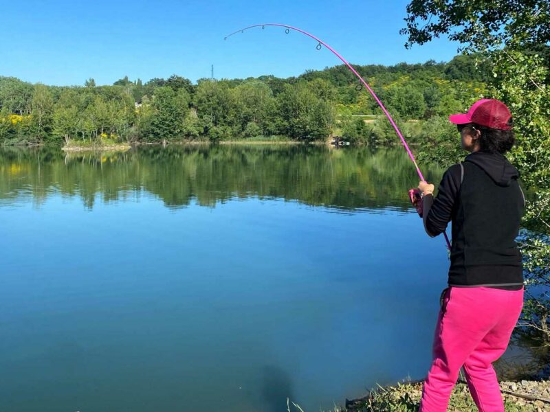 Female angler in pink trousers and a pink cap playing a fish, her pink fishing rod bent right over, by a lake