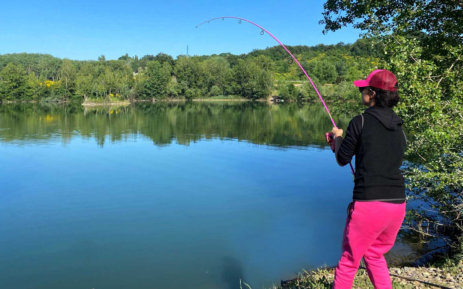 Anglerin in pinker Hose und pinker Cap im Drill mit stark gebogener pinker Angelrute am See