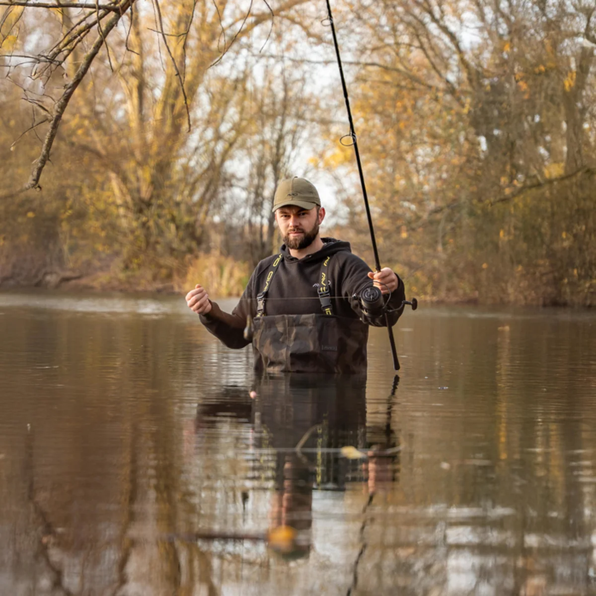 Ribič u Avid Carp ribolovnim čizmama-hlačama sa šaranskim štapom stoji u vodi ispred jesenskog drveća