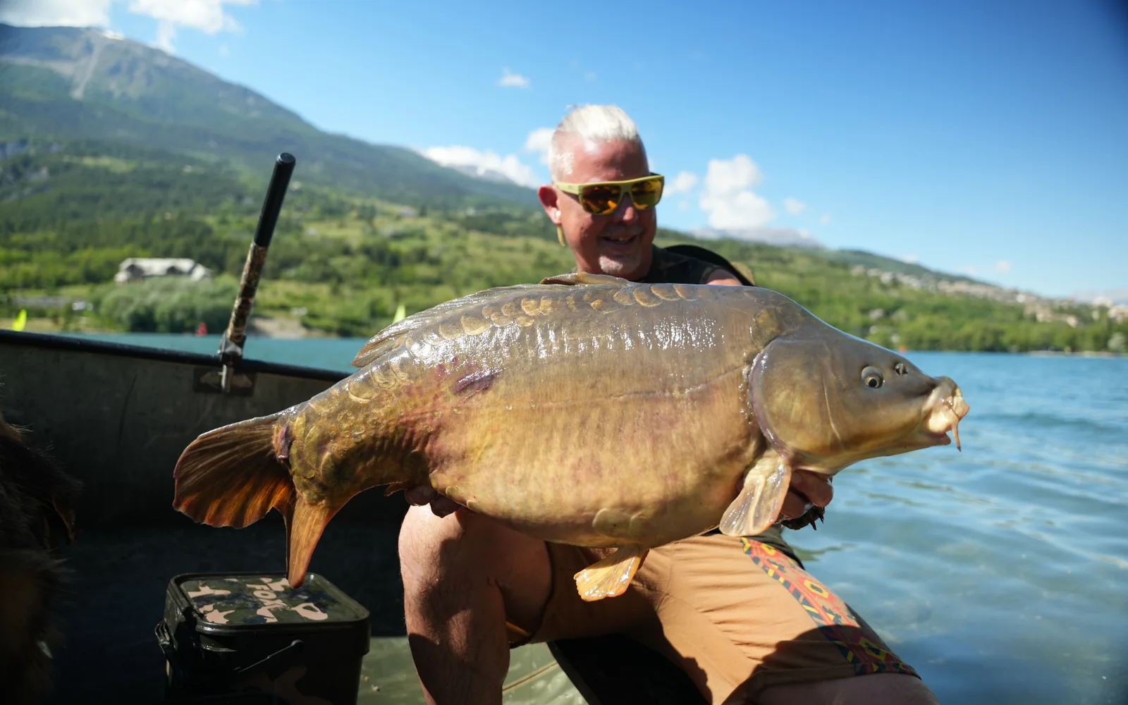 Angler holding a big mirror carp in a boat with mountains in the background.